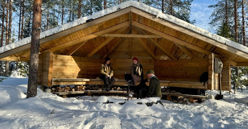 Kolme ihmistä ja koira istuvat LockCornerin tekemällä laavulla talvisissa maisemissa.