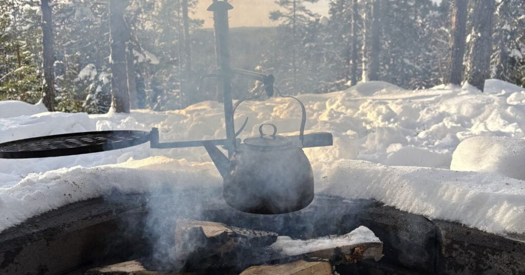 Kuvassa laavulla talvisessa maisemassa nuotio, jossa keitetään nokipannukahvia.