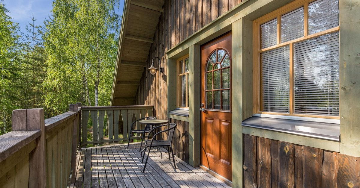Hirsirakennus, jossa ruskea ovi ja vihreät ikkunapuitteet. Taustalla sininen taivas ja koivuja.