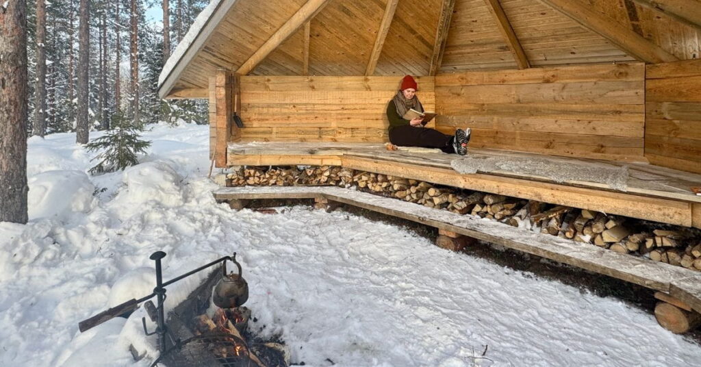 Nainen lukee kirjaa laavulla. Laavun edessä tulipaikka jossa kahvipannu.
