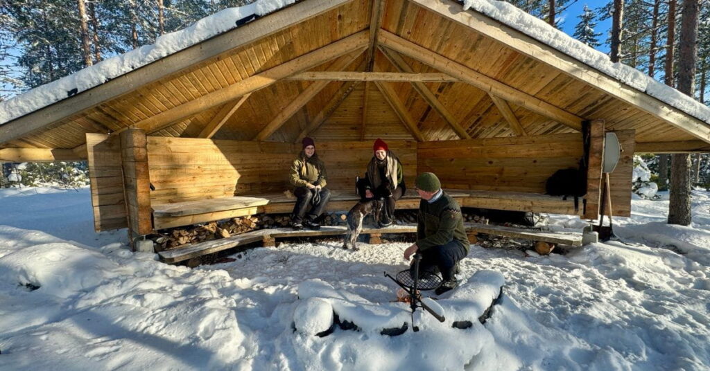 Kuvassa laavulla ihmisiä. Laavun edessä tulipaikka, jossa mies sytyttää tulta.