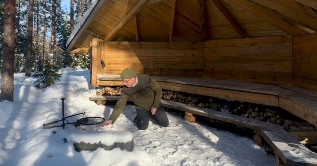 Mies on laavun edessä tulipaikalla sytyttämässä tulta. Talvinen päivä.