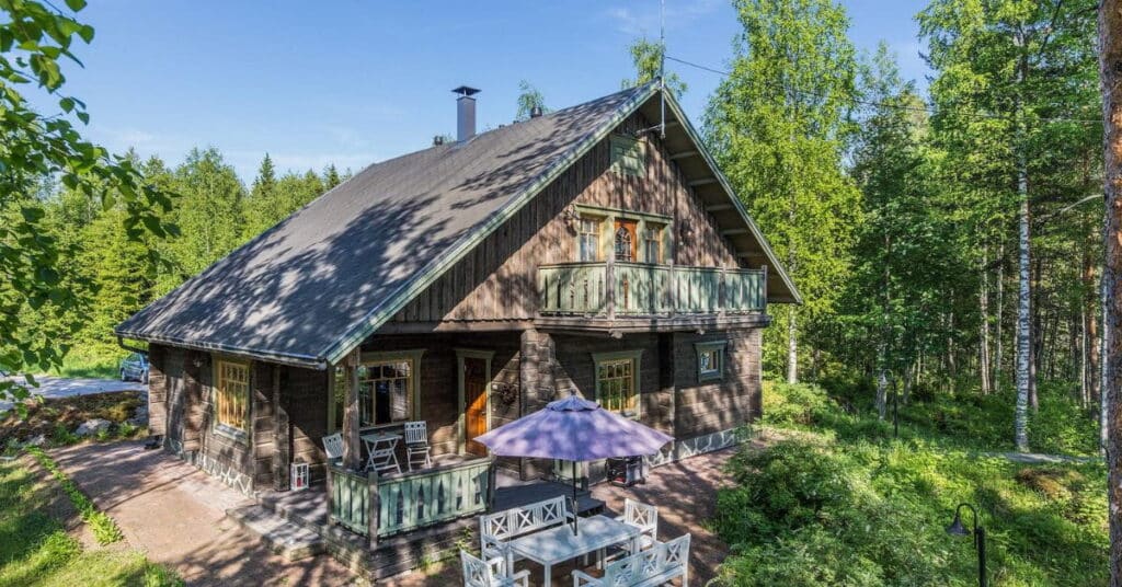 Kaksikerroksinen hirsitalo metsän keskellä, edessä istuinryhmä.